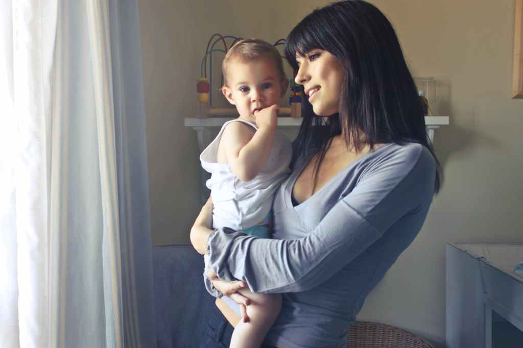 woman carrying baby boy wearing white tank top infront of white curtain inside the room